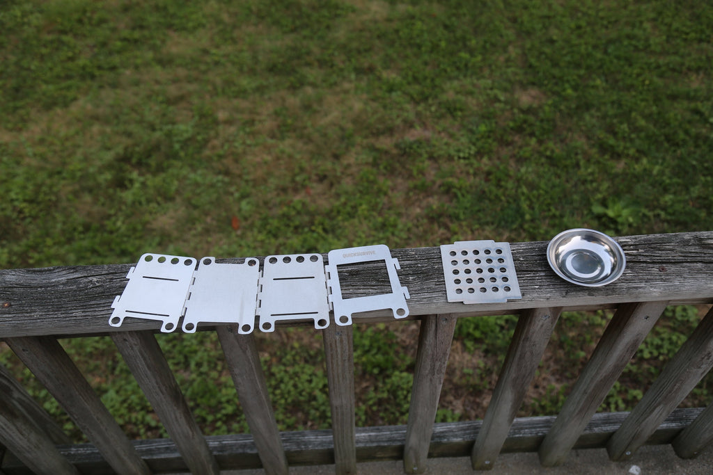 The components of the Portable Mini Foldable Wood Burning Survival Stove by QUICKSURVIVE and a small round dish are neatly arranged on a wooden railing, with grass in the background, ready to be packed into your survival packs for your next adventure.