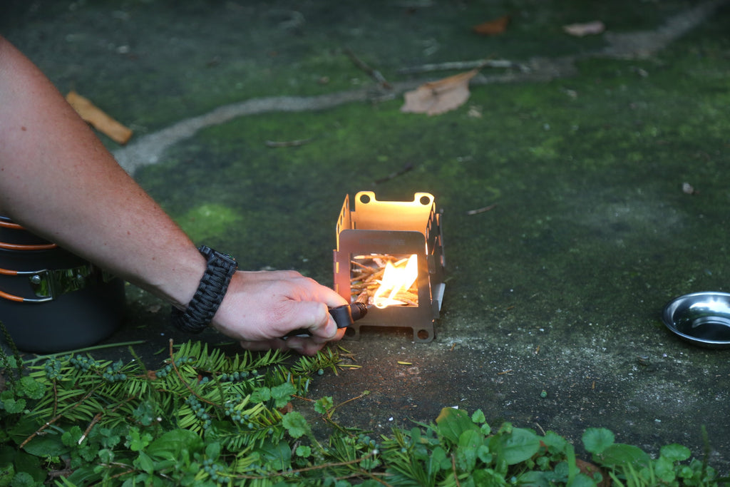 A person uses a fire starter to ignite a Portable Mini Foldable Wood Burning Survival Stove by QUICKSURVIVE on a mossy concrete surface outdoors, with camping equipment and survival packs visible nearby.