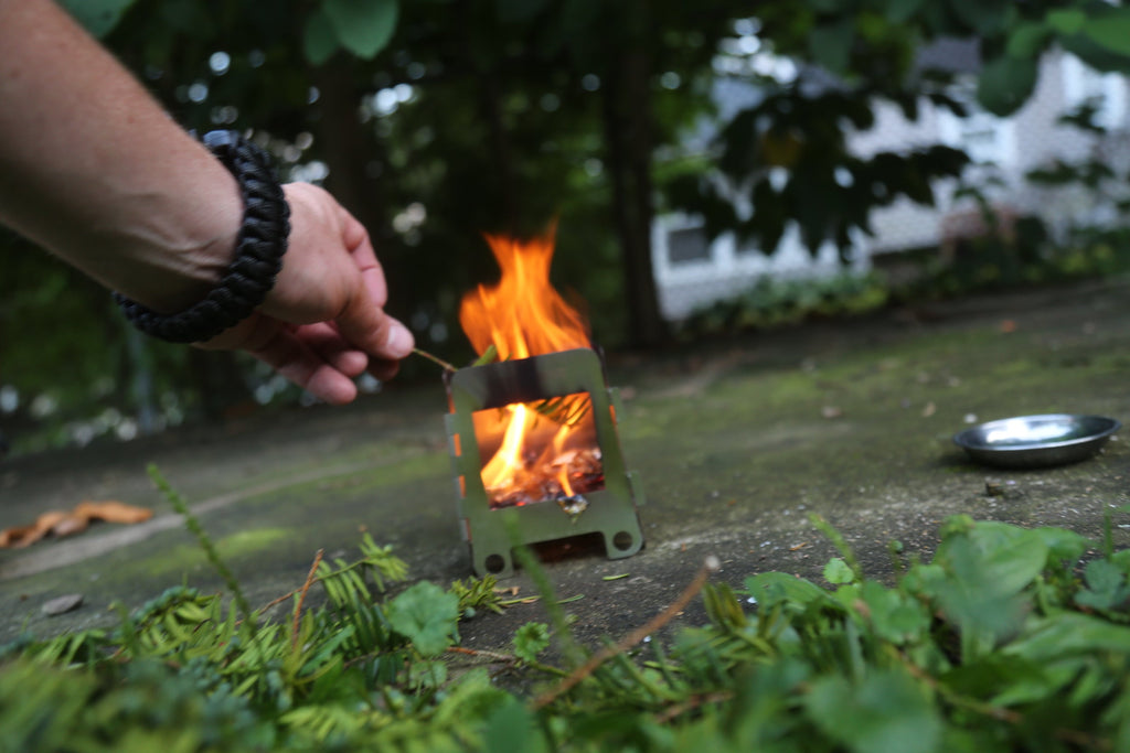 A person is igniting a Portable Mini Foldable Wood Burning Survival Stove by QUICKSURVIVE in a lush garden area, foliage enveloping the scene, epitomizing the convenience of QUICKSURVIVE's portable camping stoves often found in survival packs.