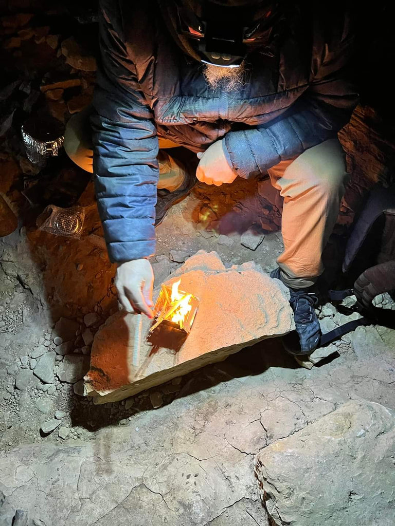 A person crouches on rocky ground, using a headlamp and igniting a small fire on a flat rock with the compact efficiency of the Portable Mini Foldable Wood Burning Survival Stove by QUICKSURVIVE.