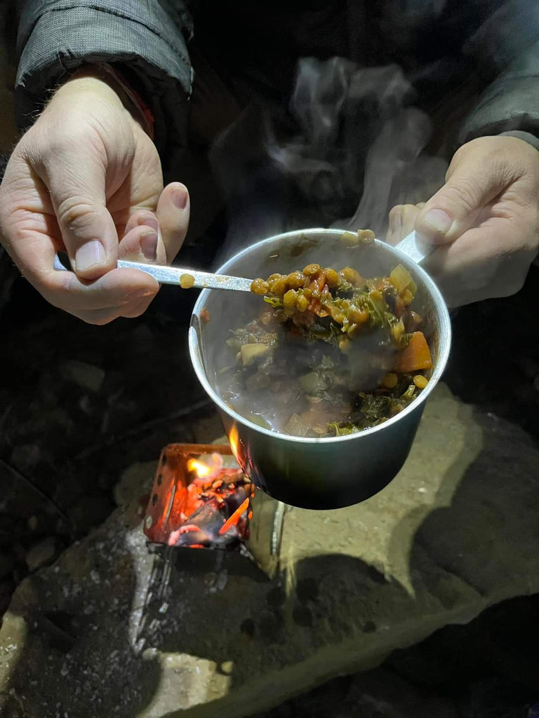 A person stirs a pot of vegetable stew over a small open flame in an outdoor setting, using the reliable Portable Mini Foldable Wood Burning Survival Stove by QUICKSURVIVE.