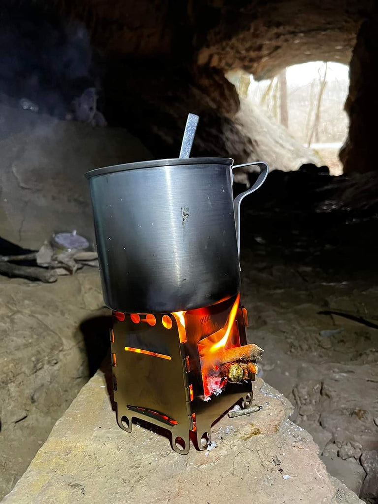 A metal pot with a spoon inside is placed on a Portable Mini Foldable Wood Burning Survival Stove by QUICKSURVIVE, emitting smoke inside a cave. An opening to the outside is visible in the background, hinting at the necessity of QUICKSURVIVE survival packs nearby.