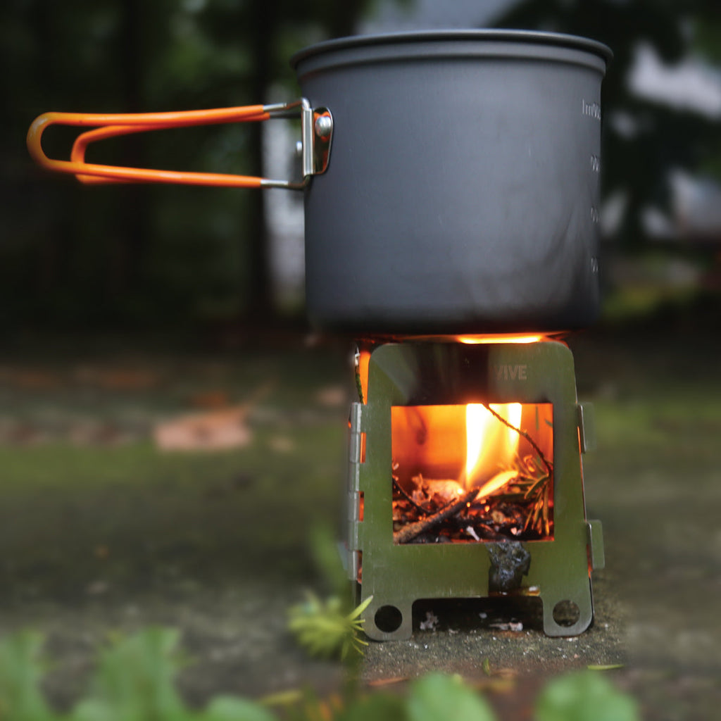 A black pot with orange handles is placed on a small Portable Mini Foldable Wood Burning Survival Stove by QUICKSURVIVE, its flames crackling warmly in the outdoors, making it perfect for survival packs.