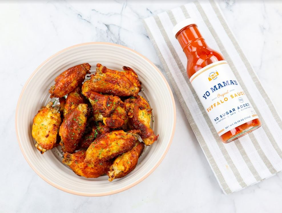 A bowl of Grilling Keto Condiment Gift Set (Keto and Paleo Certified) chicken wings next to a bottle of Yo Mama's Foods sauce, perfect for Foodies.
