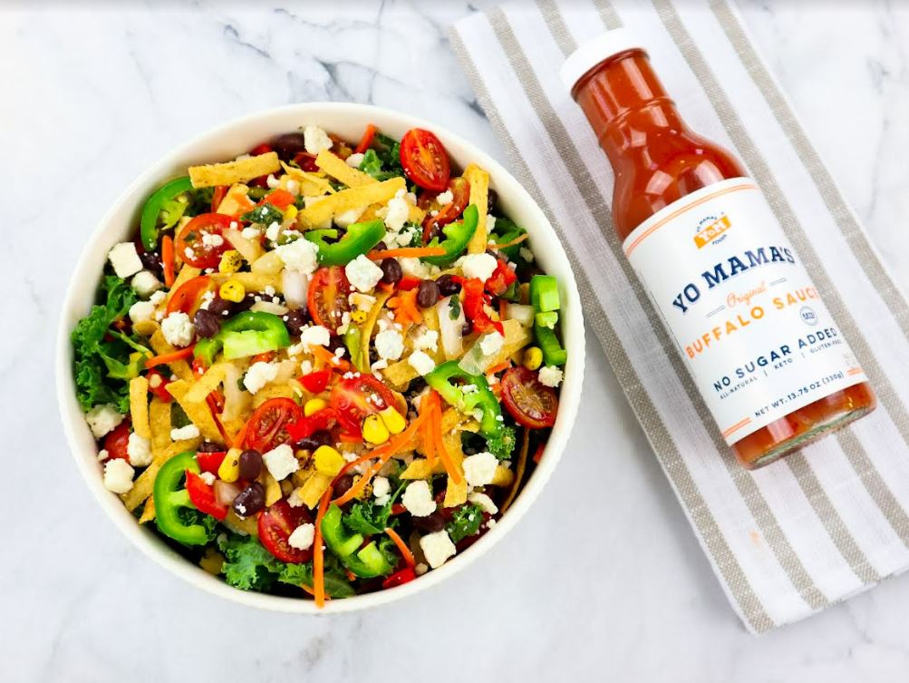 A bowl of salad with a bottle of Classic Buffalo sauce by Yo Mama's Foods next to it, perfect for XSpecial Marketplace foodies.