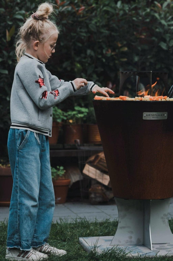 A little girl is standing in front of an Arteflame Outdoor Grills One Series 20" Grill, appealing to foodies.