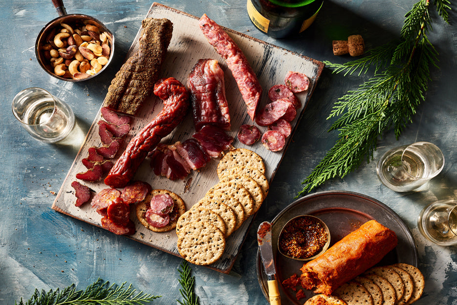 A XSpecial Marketplace platter of Classic Box by Carnivore Club USA, featuring meat, cheese, crackers and wine on a blue background.